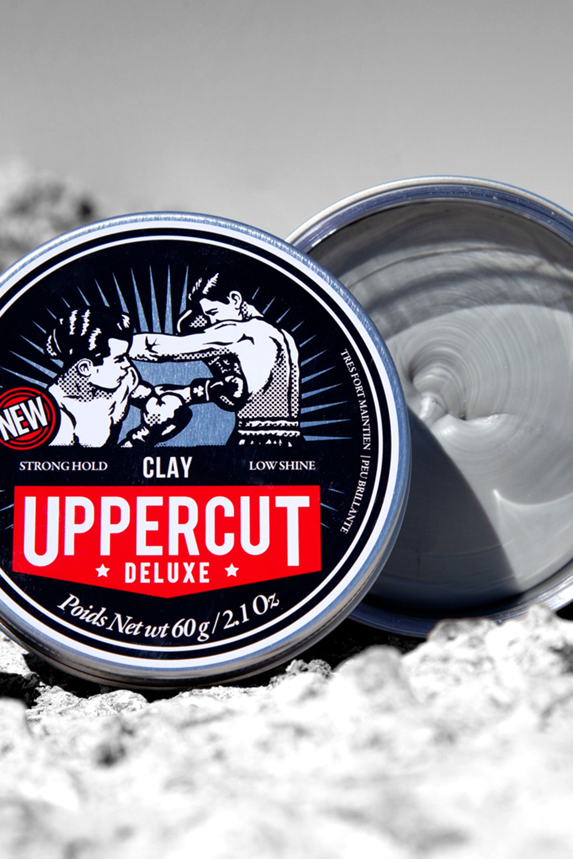 An open tin of Other Uppercut Deluxe Clay sits on a rocky surface, revealing gray clay. The label, with two vintage-style boxers, highlights its strong hold, matte finish, and low shine.