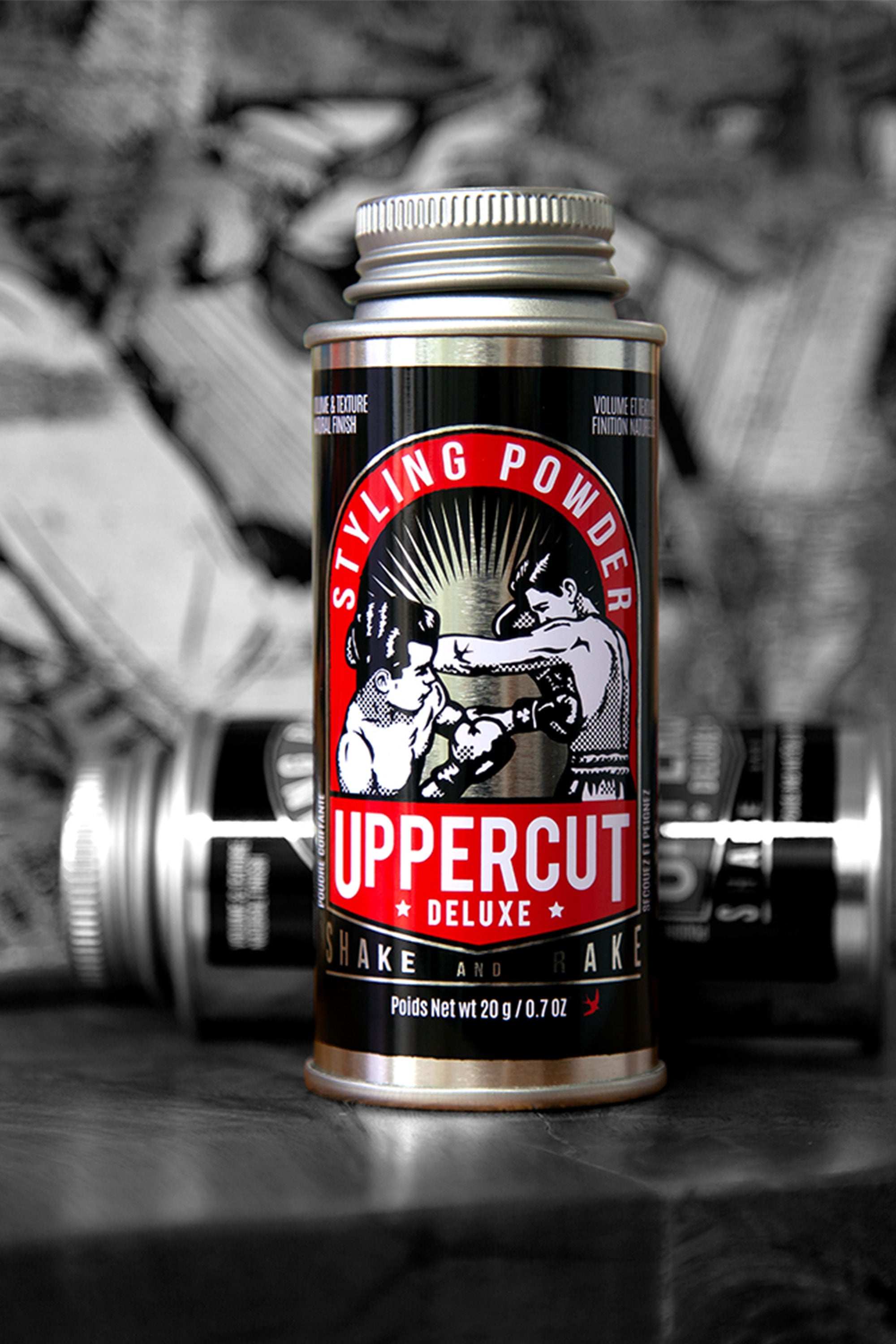 A 20g canister of Uppercut Styling Powder by Other stands upright, featuring bold red, black, and white branding with a vintage boxer illustration. Blurred cans appear in the monochrome background, highlighting its matte finish and volumizing effect.