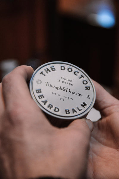 A person’s hands holding a round tin of Triumph & Disaster’s The Doctor – Beard Balm. Its minimalist black-on-white design nods to W.G. Grace's cricket era.