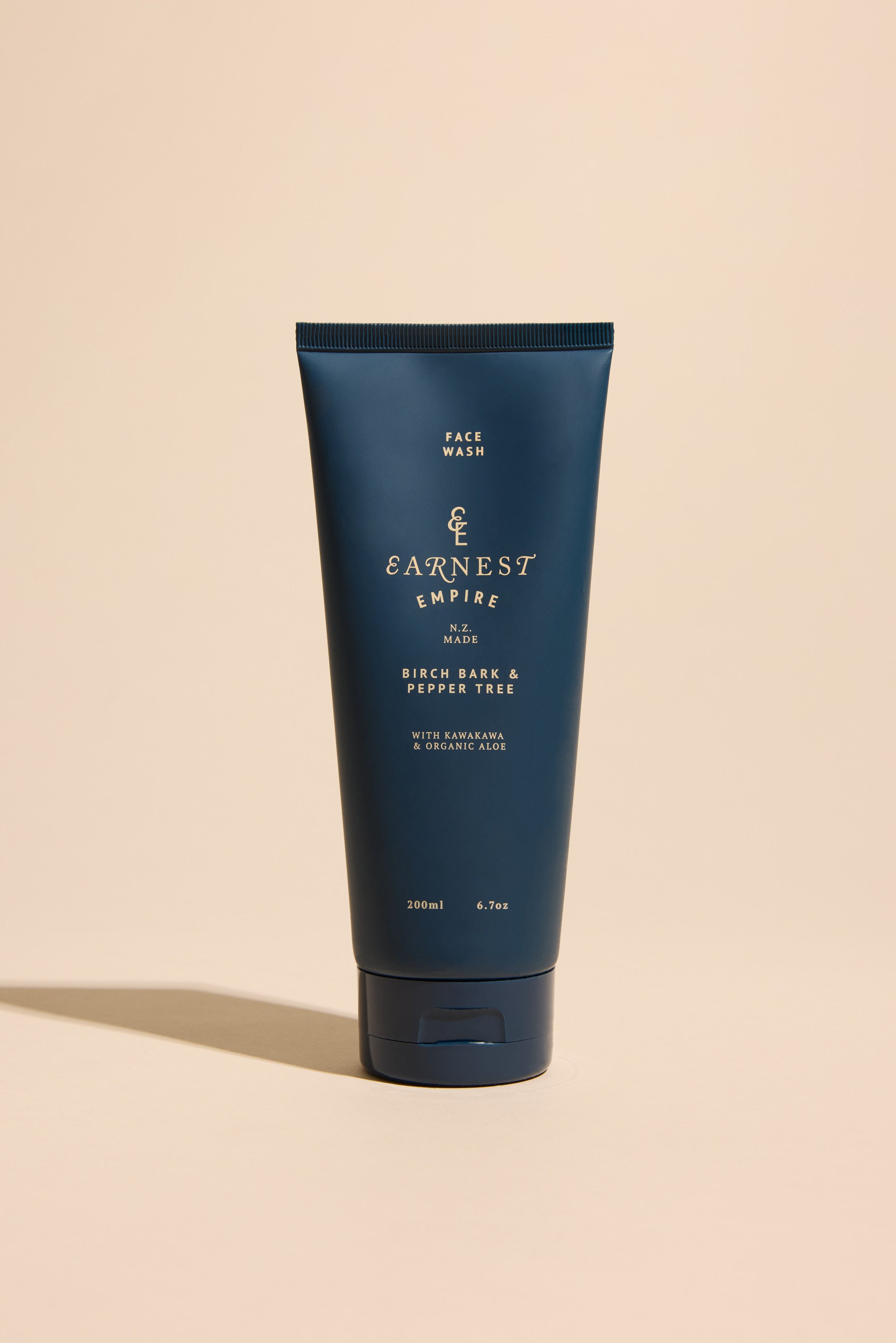 A dark blue tube of Other Earnest Empire Face Wash stands upright on a beige surface, featuring gold and white text that highlights its natural cleansing formula and size (200ml/6.7oz).