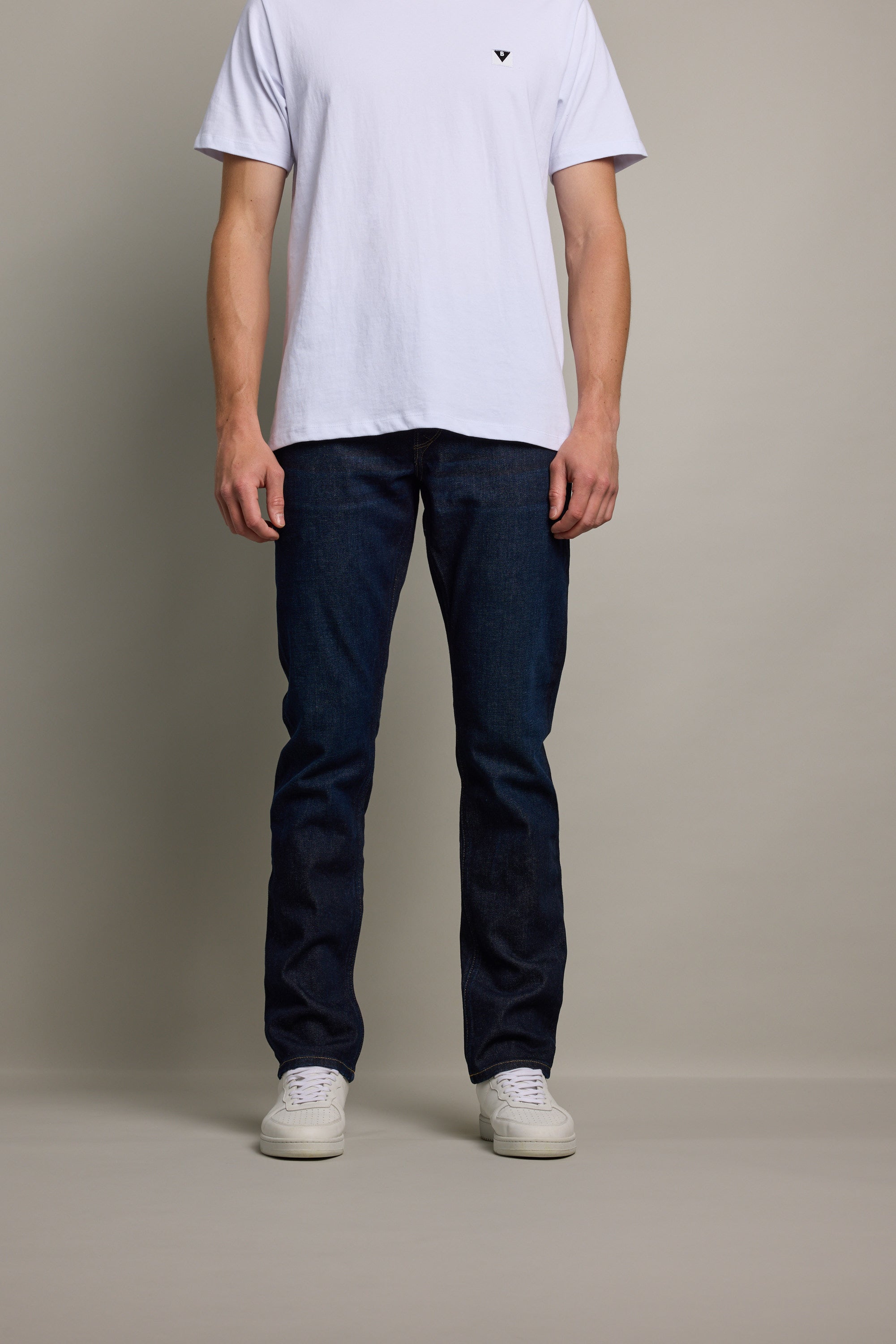 A person stands against a plain background wearing a white short-sleeve tee, white sneakers, and Barkers Nomad Denim Taper Jean in an athletic fit. The sustainable denim jeans are featured prominently; the person's face is not visible.