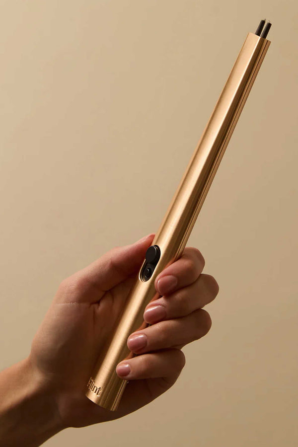 A hand holds the Flint Rechargeable Lighter 2.0 by Flint, a slim metallic gold lighter with a black ignition button, against a plain beige background.