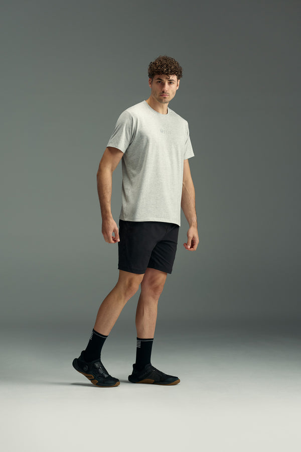 A man with curly hair stands on a plain backdrop, wearing a light gray t-shirt and black WearEver Agile Short by WearEver with an elastic waistband, paired with black socks and running shoes. He faces slightly to the side with a neutral expression.