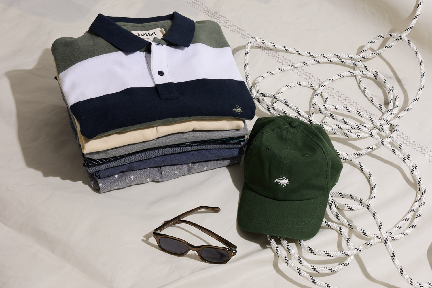 A folded stack of shirts, a green baseball cap with a white palm tree logo, a pair of sunglasses, and a coiled rope are arranged on a light-colored surface.