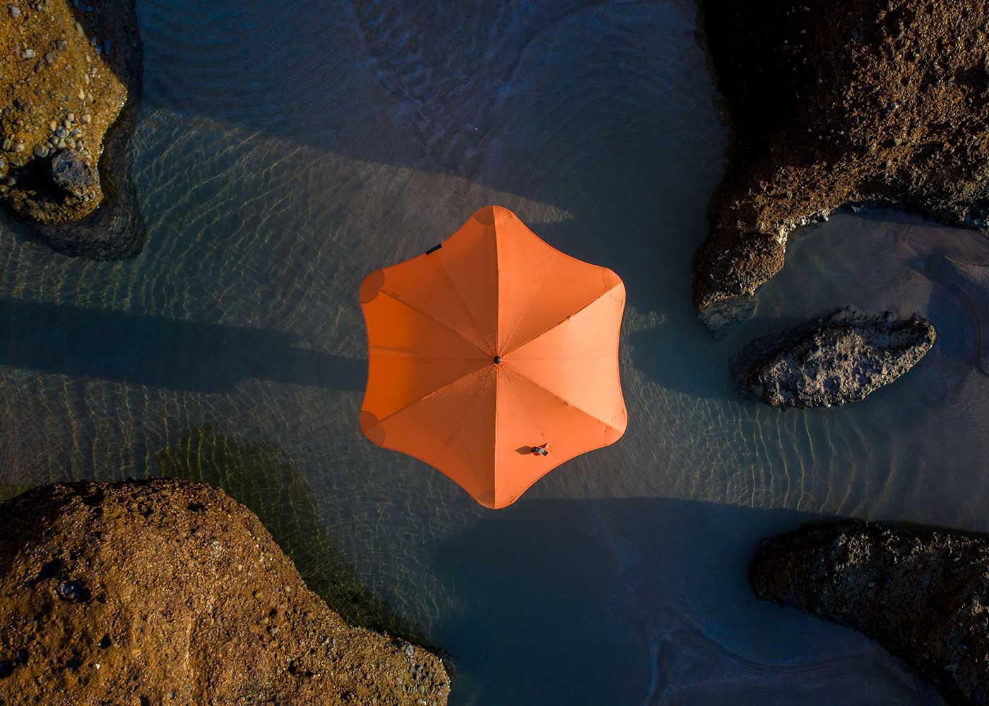 A bright orange umbrella viewed from above sits on shallow water, surrounded by rocks and casting a shadow in the rippling blue water.