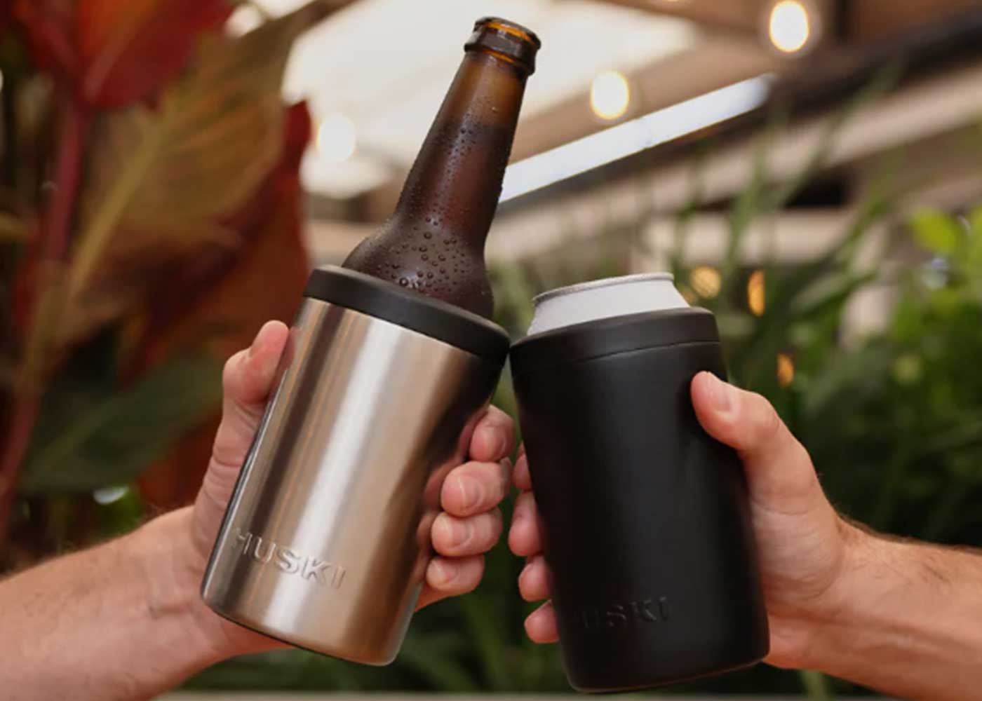 Two hands toast with drinks, one holding a brown bottle in a silver Huski cooler, the other holding a can in a black Huski cooler, against a blurred, leafy background.