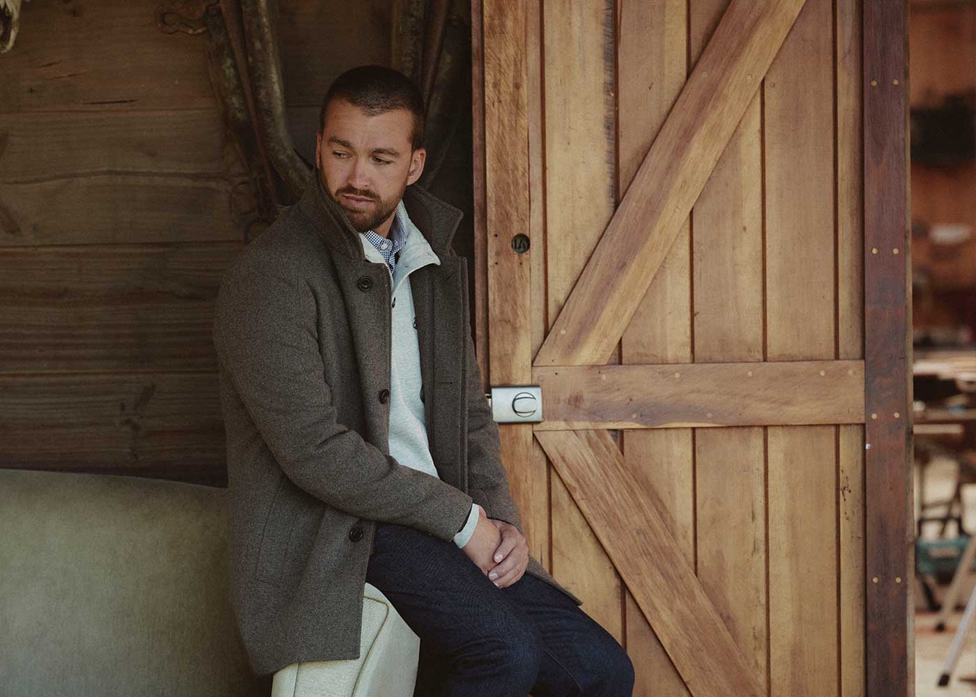 A man with a short beard sits on a bench near a wooden barn door, wearing a gray coat over a light sweater and dark jeans, looking to his left with a thoughtful expression.