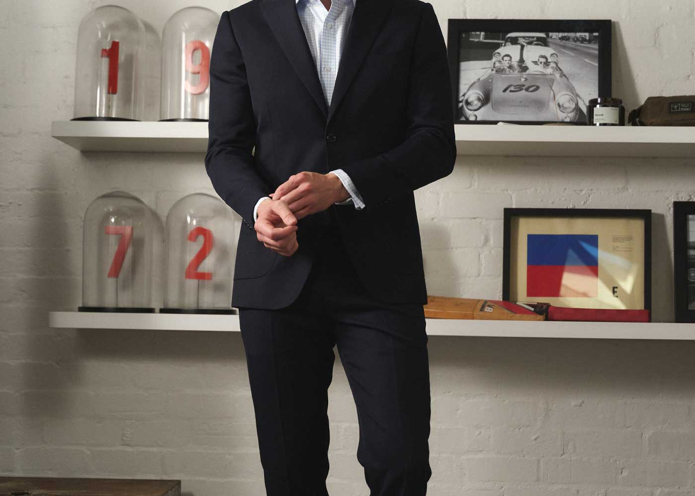 A person in a dark suit adjusts their cuff in a modern room with white shelves holding glass domes labeled 1, 9, 7, 2, and framed art and photos on a white brick wall.