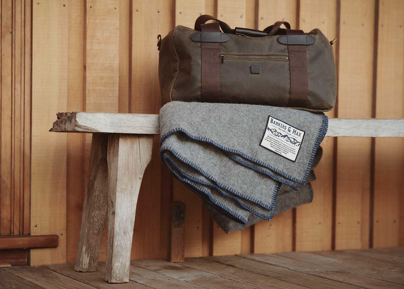 A green duffel bag with brown straps sits on a rustic wooden bench, alongside a folded gray wool blanket with blue trim and a visible label, against a wooden-paneled wall.