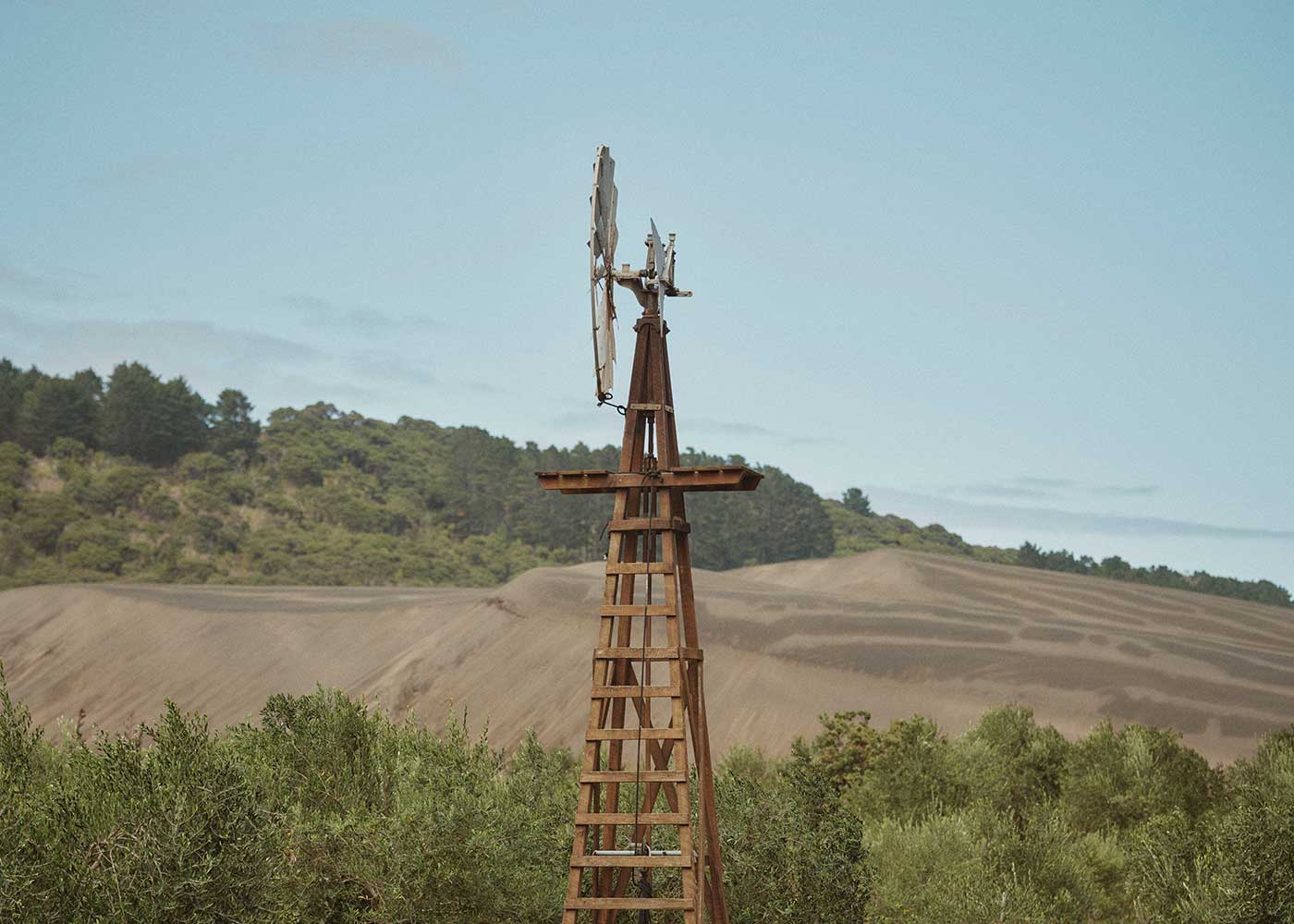 A wooden windmill stands among green shrubs with rolling hills and trees in the background under a clear blue sky.