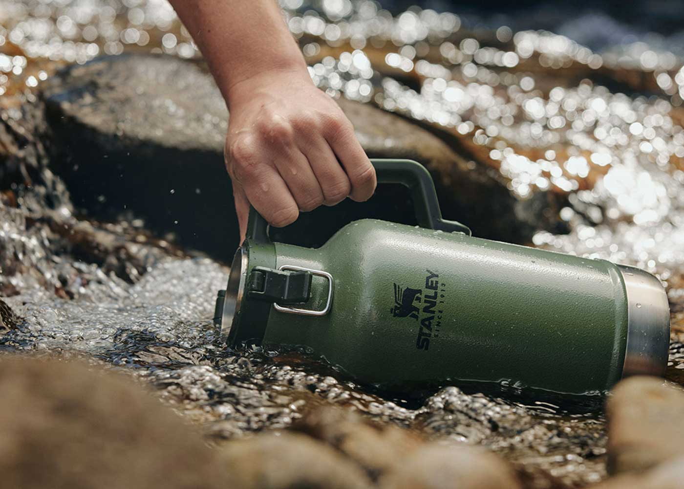 A hand holds a green Stanley water bottle in a shallow, rocky stream, filling it with water.