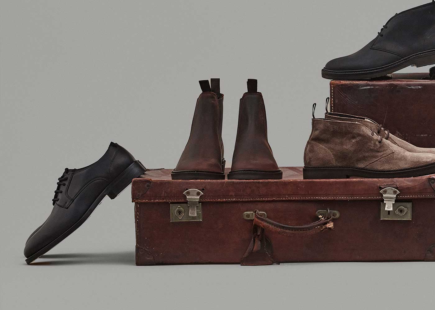 Four pairs of mens dress shoes in black, brown, and taupe are displayed on and around two stacked vintage leather suitcases against a plain gray background.