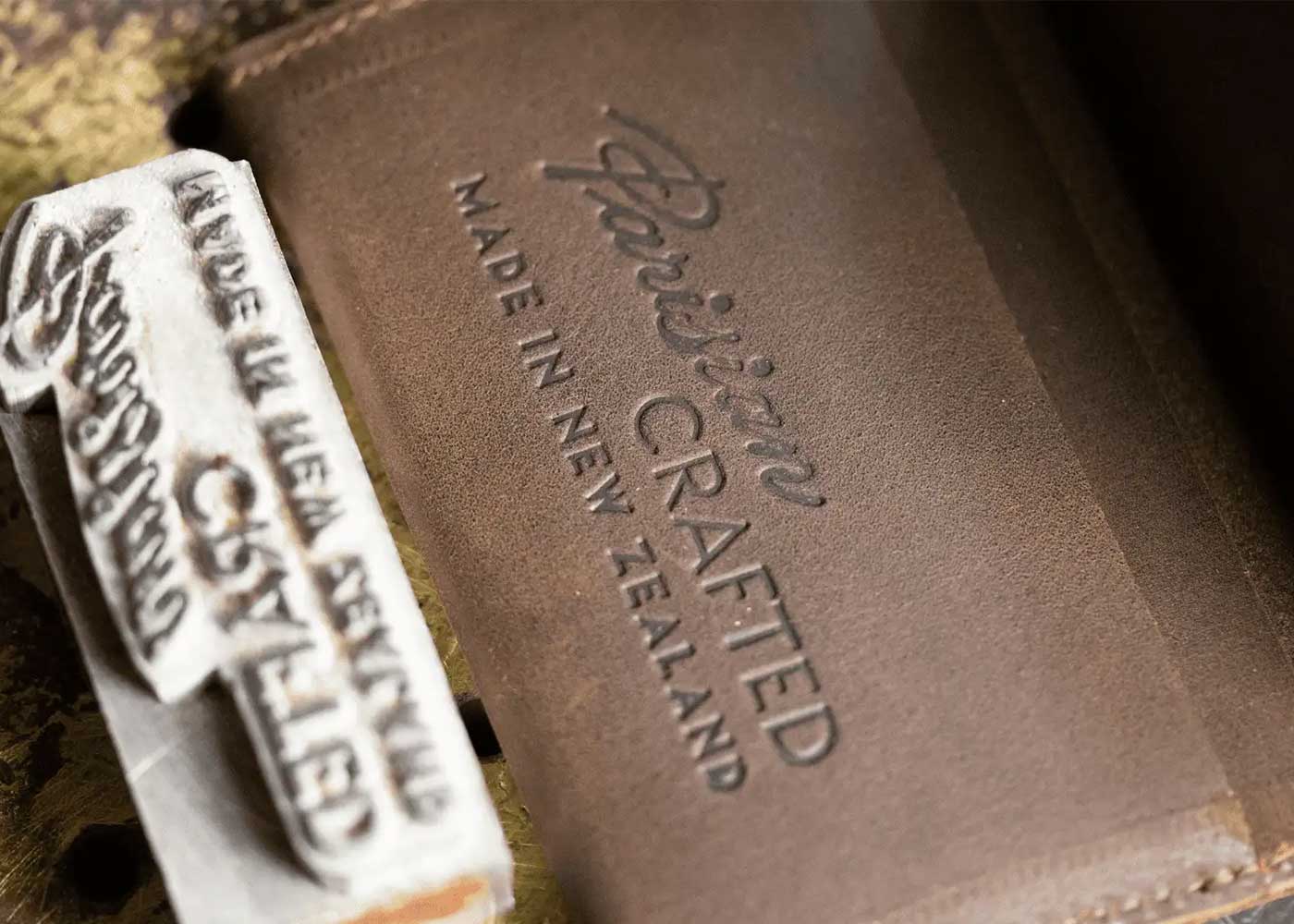 Close-up of a leather patch embossed with Parisian CRAFTED MADE IN NEW ZEALAND next to a metal stamp that reads MADE IN NEW ZEALAND CRAFTED.