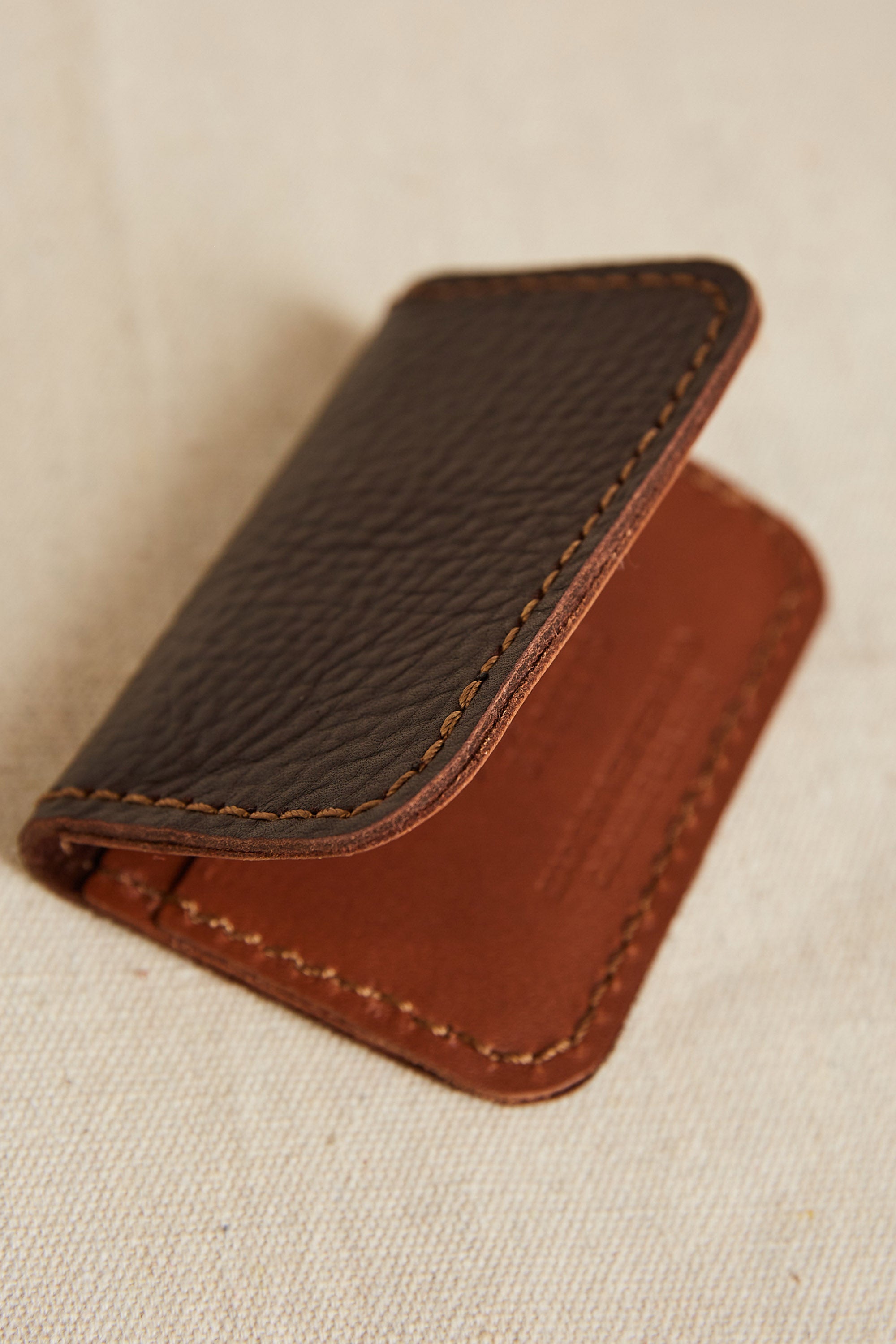 The Parisian Leather Fold Wallet by Parisian is a compact brown leather wallet with visible stitching, shown partially open on a beige fabric surface—a refined accessory inspired by Parisian style.