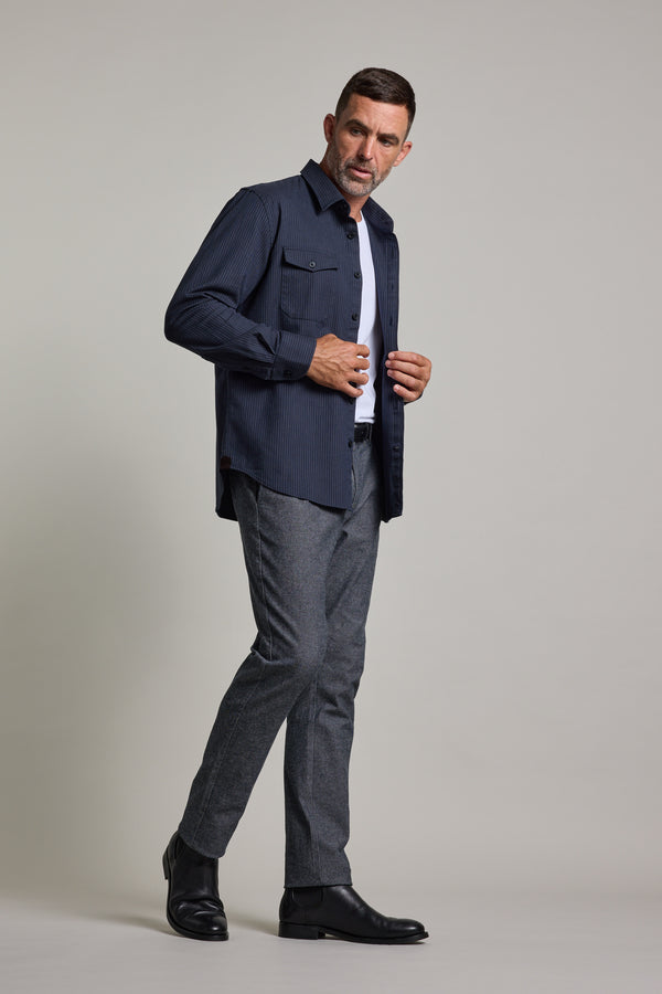 A man with short dark hair and a beard models the Barkers Timothy Stripe Overshirt over a white tee, styled with gray trousers and black boots, against a plain gray background.