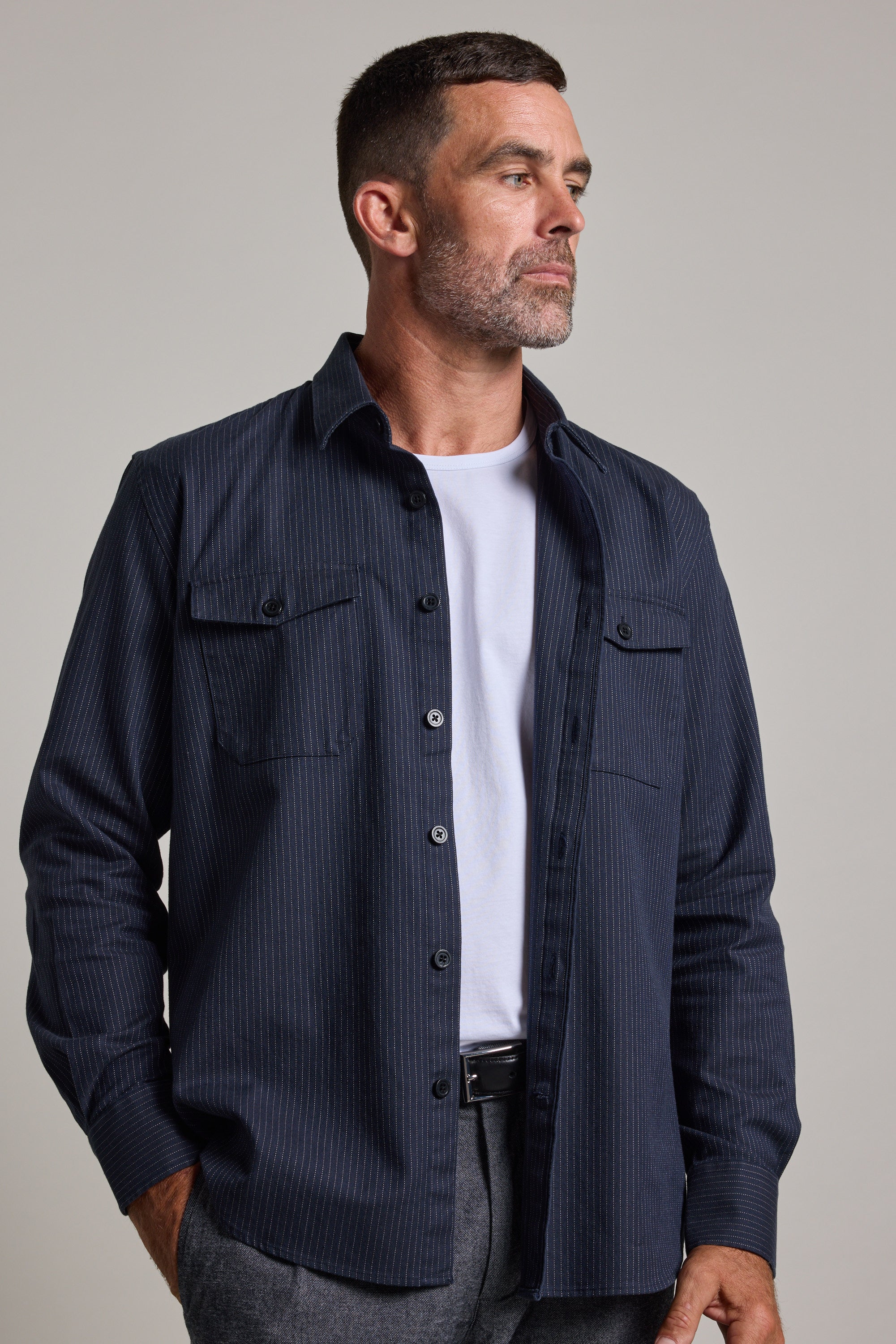 A man with short dark hair and a beard wears the Barkers Timothy Stripe Overshirt, a navy blue cotton pinstripe shirt, over a white t-shirt. He stands with one hand in his pocket, looking to the side against a plain background.