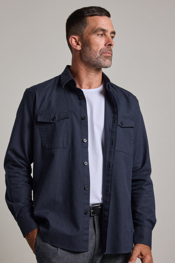 A man with short dark hair and a beard wears the Barkers Timothy Stripe Overshirt, a navy blue cotton pinstripe shirt, over a white t-shirt. He stands with one hand in his pocket, looking to the side against a plain background.