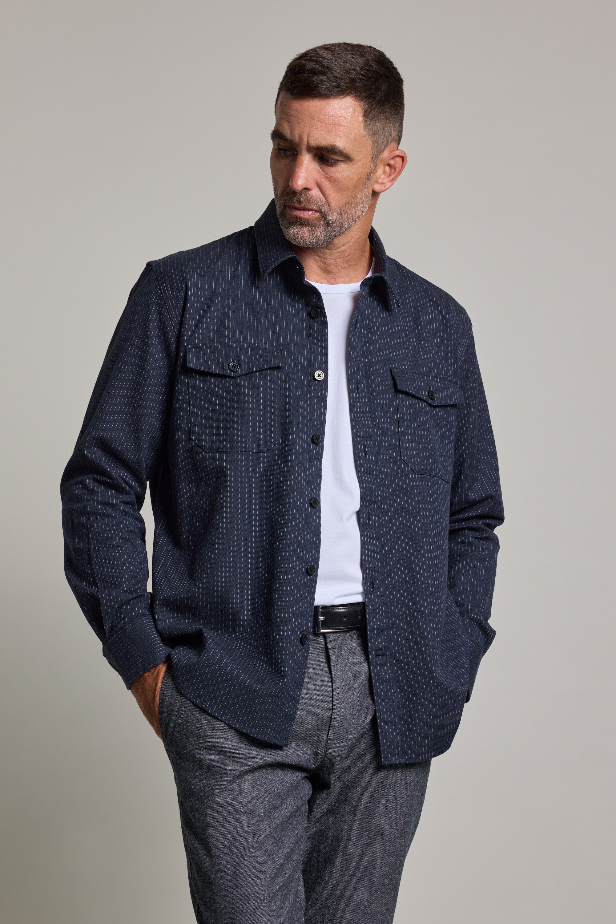 A man with short hair and a beard wears the Barkers Timothy Stripe Overshirt in navy blue over a white t-shirt and gray trousers, standing with hands in pockets and looking left against a neutral background.