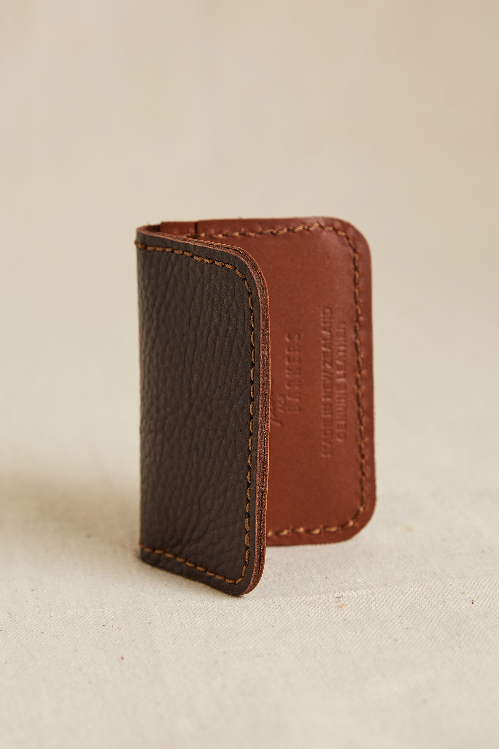 The Parisian Leather Fold Wallet by Parisian, made from New Zealand leather, has textured dark brown leather on one side and smooth light brown on the other, and stands upright on a neutral fabric surface.
