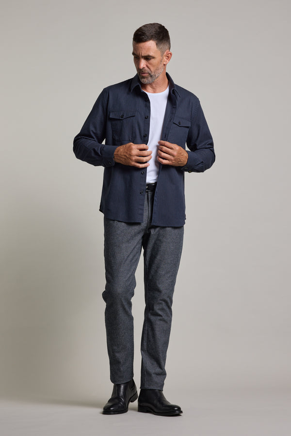 A man with short dark hair and a trimmed beard models the Barkers Timothy Stripe Overshirt over a white t-shirt, styled with gray pants and black boots, standing against a plain light backdrop and looking slightly left.