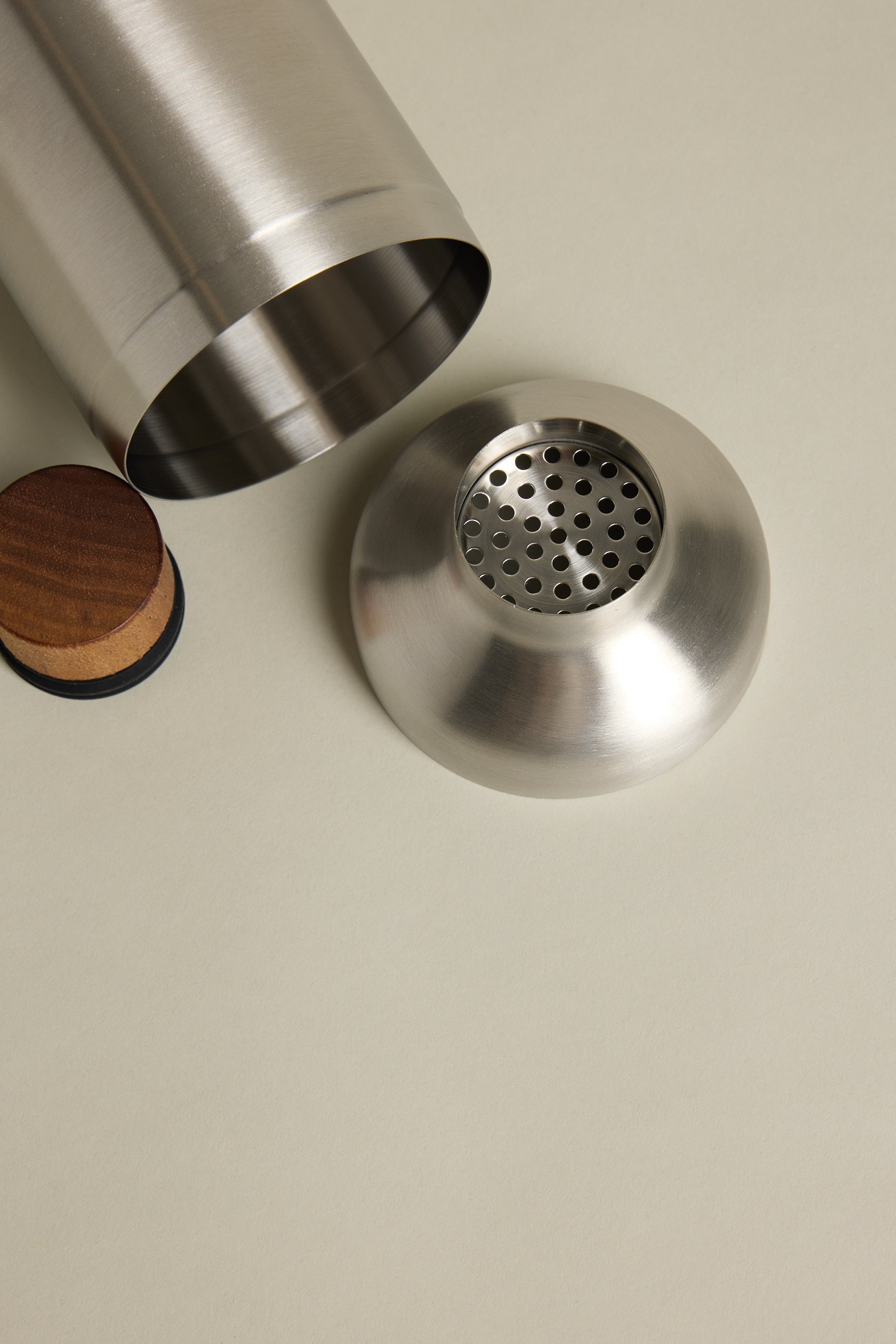 The Other Liquid Lounge Cocktail Shaker, with its lid and strainer removed, rests on a light beige surface beside a round wooden coaster.