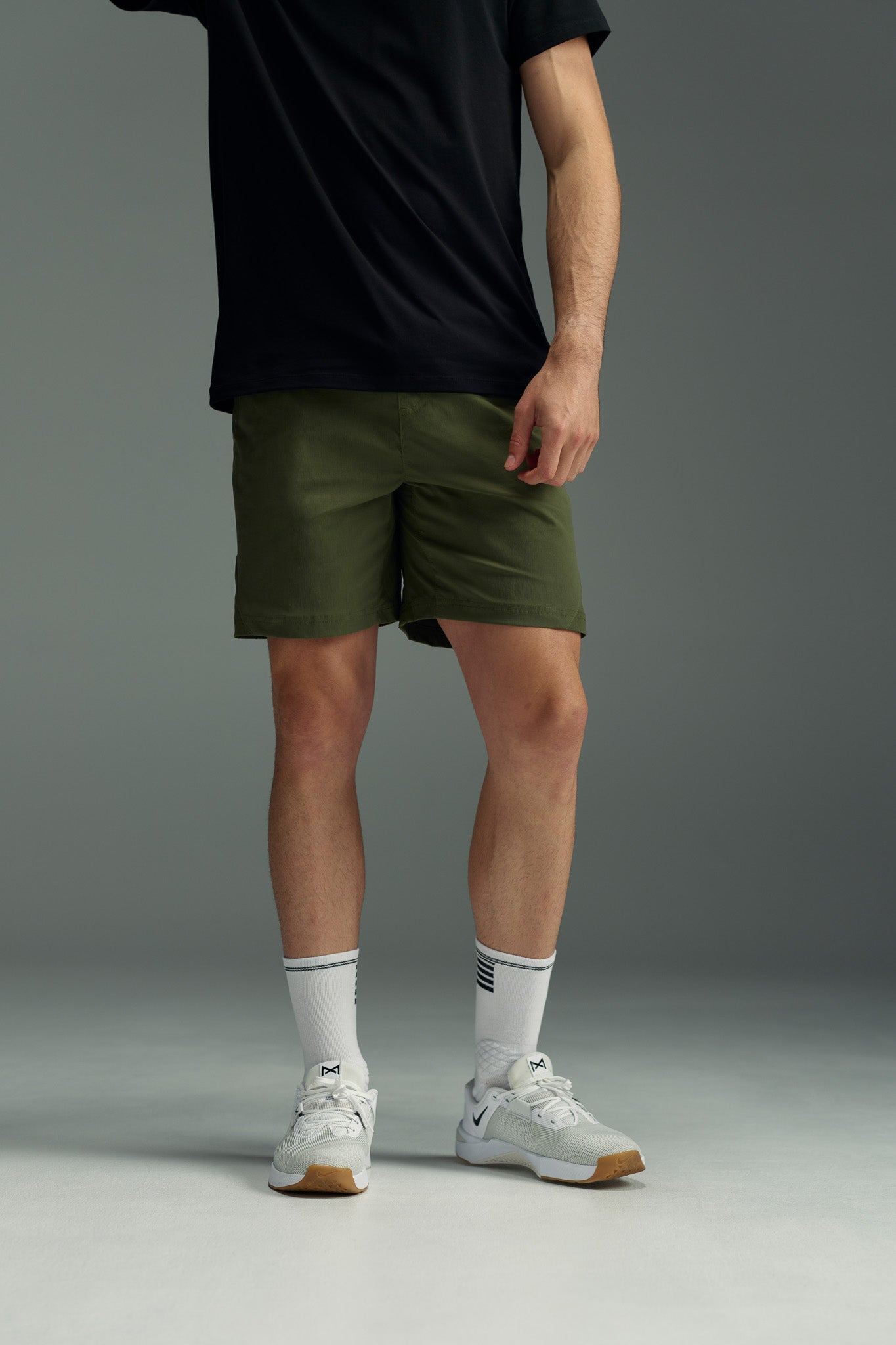 A person stands against a plain gray background wearing an untucked black t-shirt, WearEver Agile Short in olive green with a 7-inch inseam, white athletic socks, and white sneakers. Their head is not visible in the image.