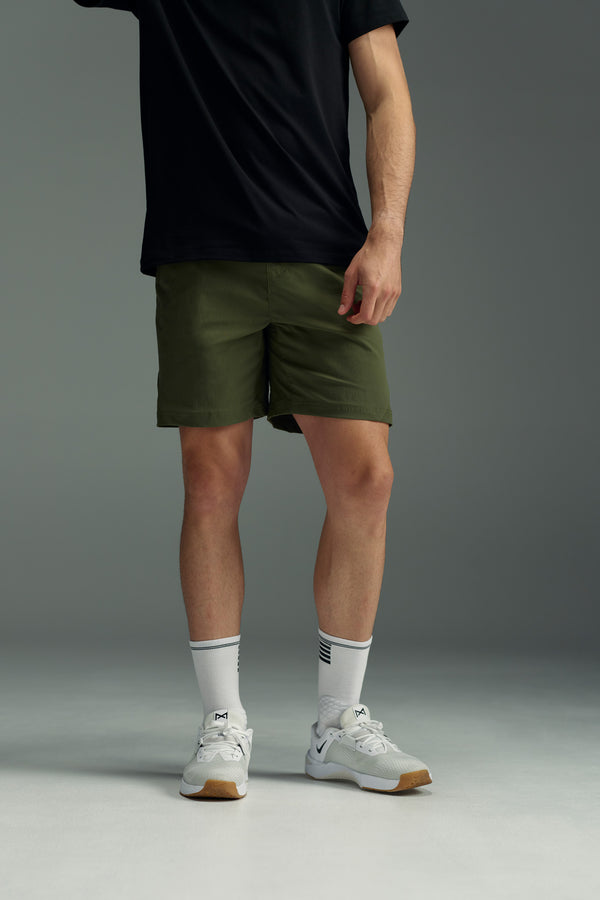 A person stands against a plain gray background wearing an untucked black t-shirt, WearEver Agile Short in olive green with a 7-inch inseam, white athletic socks, and white sneakers. Their head is not visible in the image.