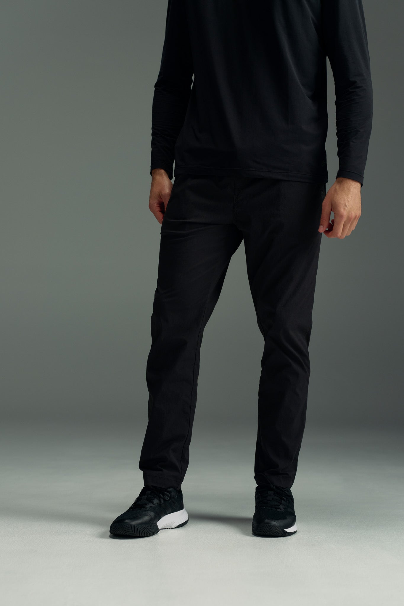 A person stands on a gray floor wearing a long-sleeve black shirt, WearEver Transit Pant by WearEver, and black athletic shoes. The upper part of their head is not visible in the image.