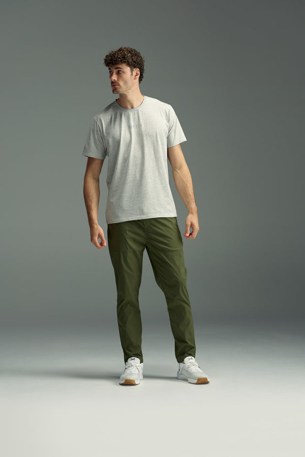 A man with short curly hair wears the WearEver Outbound T-Shirt in light gray, olive green pants, and white sneakers, standing and looking to his left against a plain gray background.