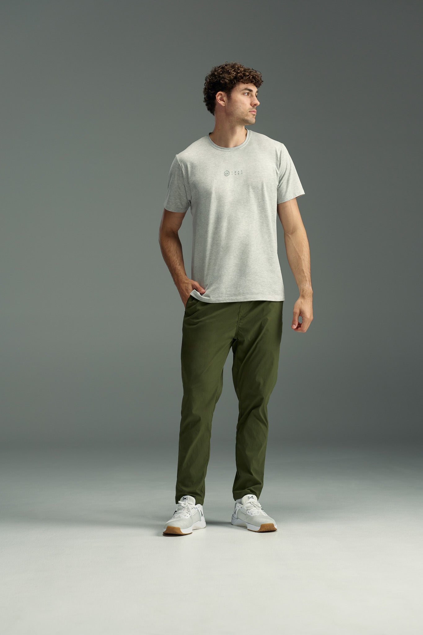 A man stands with one hand in his pocket, wearing the WearEver Outbound T-Shirt by WearEver in light gray, olive green pants, and white sneakers. He poses against a plain gray background, looking off to the side with a relaxed posture.