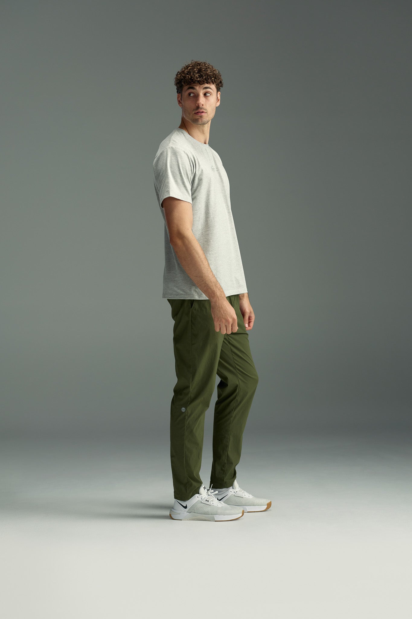 A man with curly hair stands sideways in a studio, wearing the WearEver Outbound T-Shirt by WearEver in light gray, olive green pants, and white sneakers against a plain gray background.