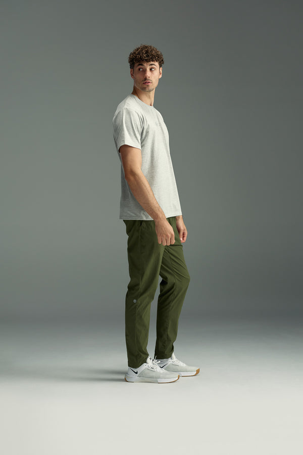 A man with curly hair stands sideways in a studio, wearing the WearEver Outbound T-Shirt by WearEver in light gray, olive green pants, and white sneakers against a plain gray background.