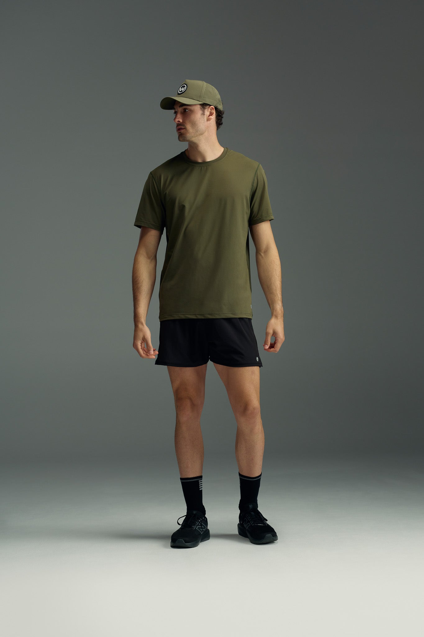A man in an olive green cap and t-shirt, black WearEver Cadence Short by WearEver, black socks, and black athletic shoes stands facing slightly left against a neutral gray background.