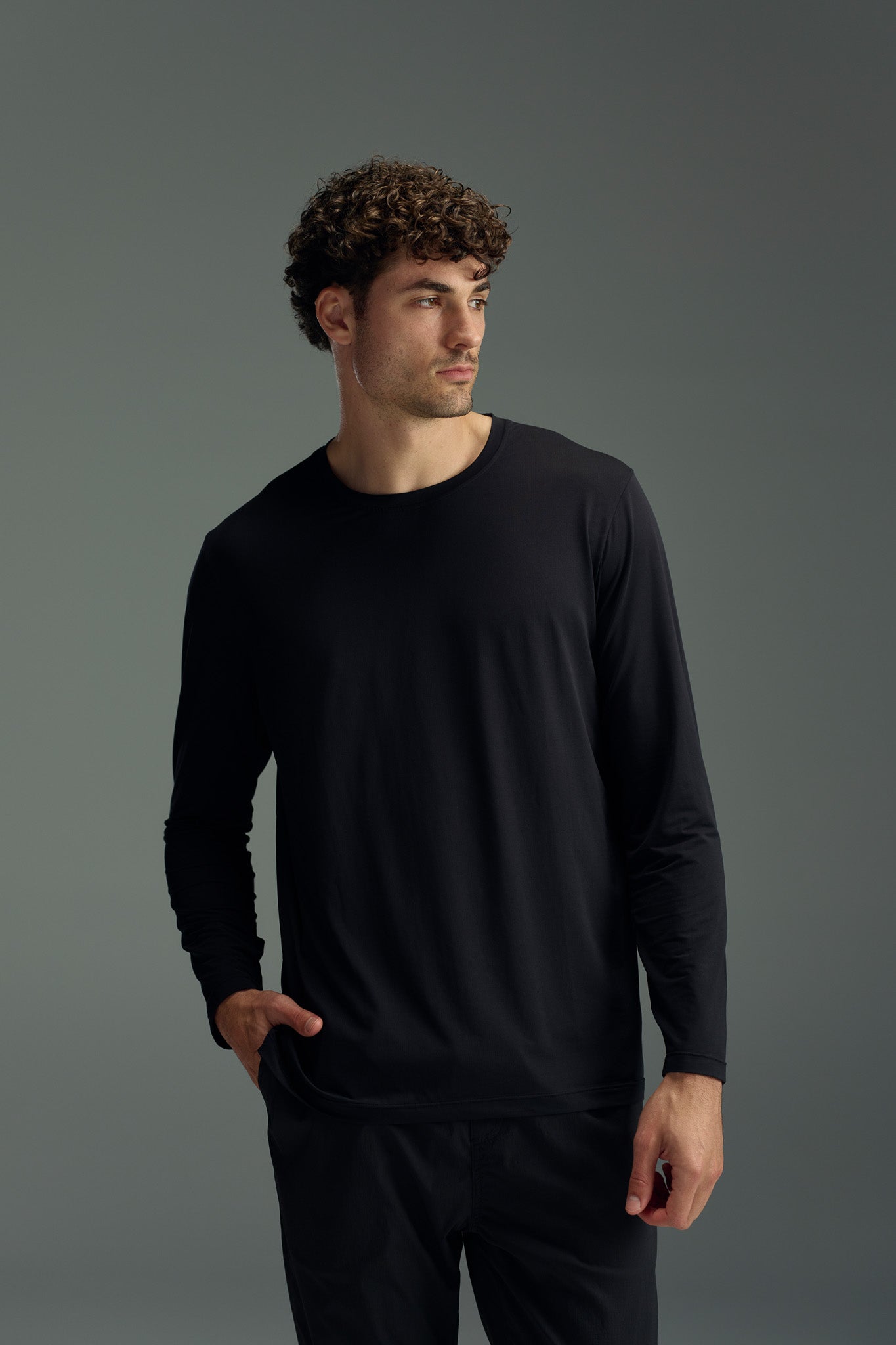 A man with curly brown hair wears the WearEver Pulse Long Sleeve Tee in black and black pants, standing against a plain gray background with one hand in his pocket, looking to the side.