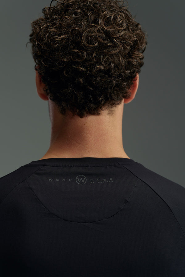 A person with short, curly brown hair is seen from behind wearing the WearEver Pulse Long Sleeve Tee in black, featuring subtle WearEver branding near the neckline against a softly lit plain background.