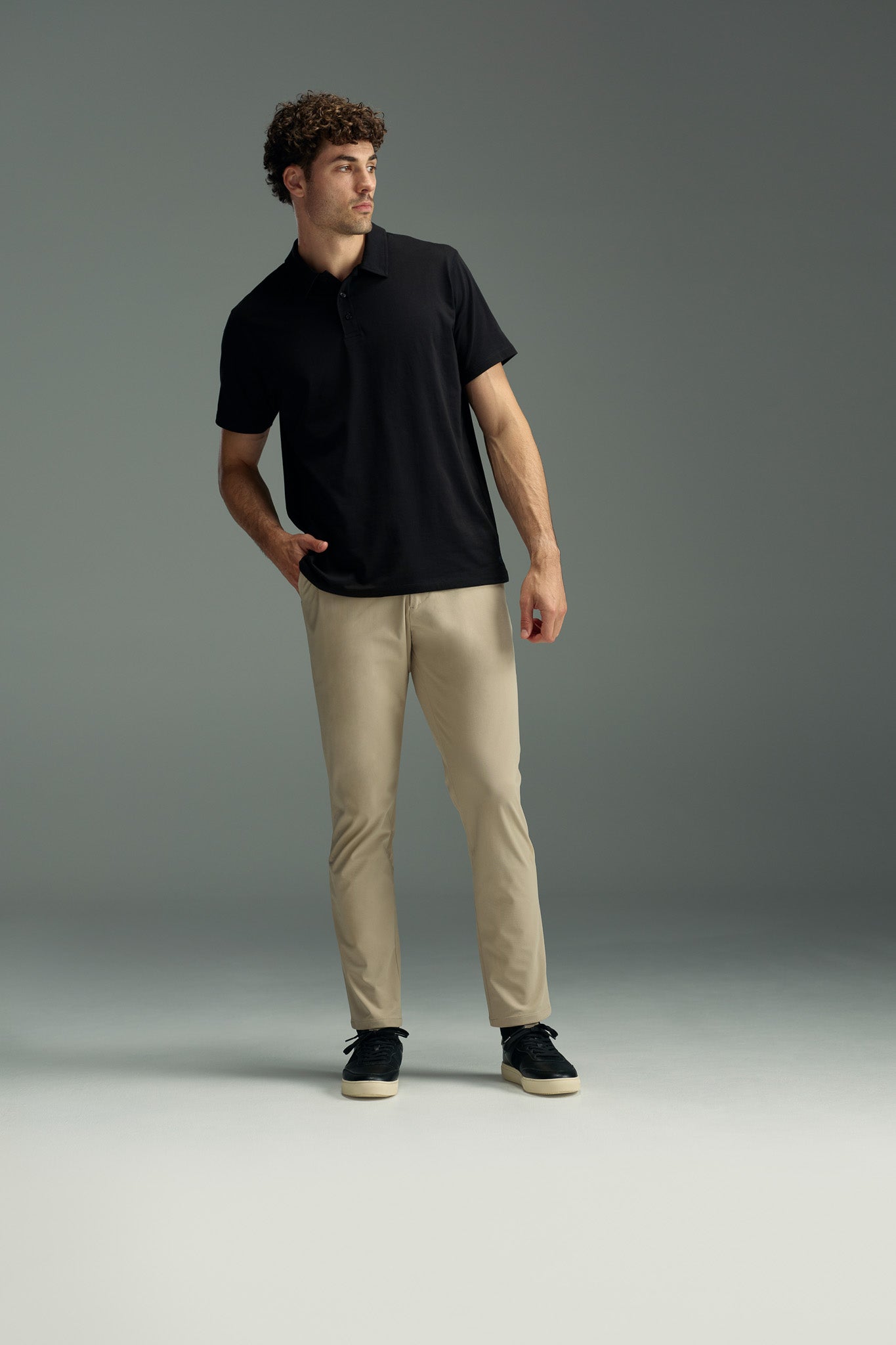 A man with curly hair stands with one hand in his pocket, looking to the side against a plain gray background. He is wearing beige WearEver Long Game Pants, a black polo shirt, and black shoes.