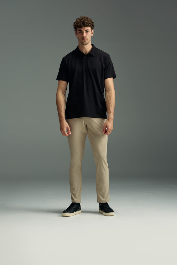 A man with curly hair faces forward, wearing a black WearEver Outbound Polo made from breathable cotton-blend fabric, beige pants, and black sneakers, standing against a plain gray background.