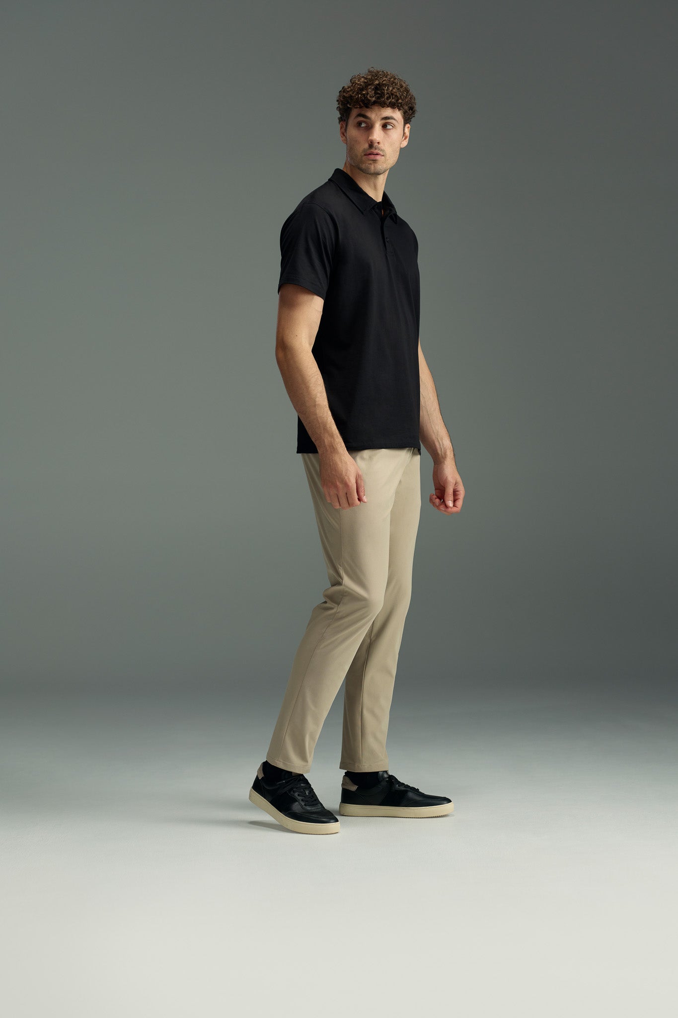 A man stands on a plain background wearing a black polo shirt and WearEver Long Game Pant by WearEver, made with lightweight, quick-dry fabric. He looks slightly to the side with one arm relaxed and the other slightly bent.