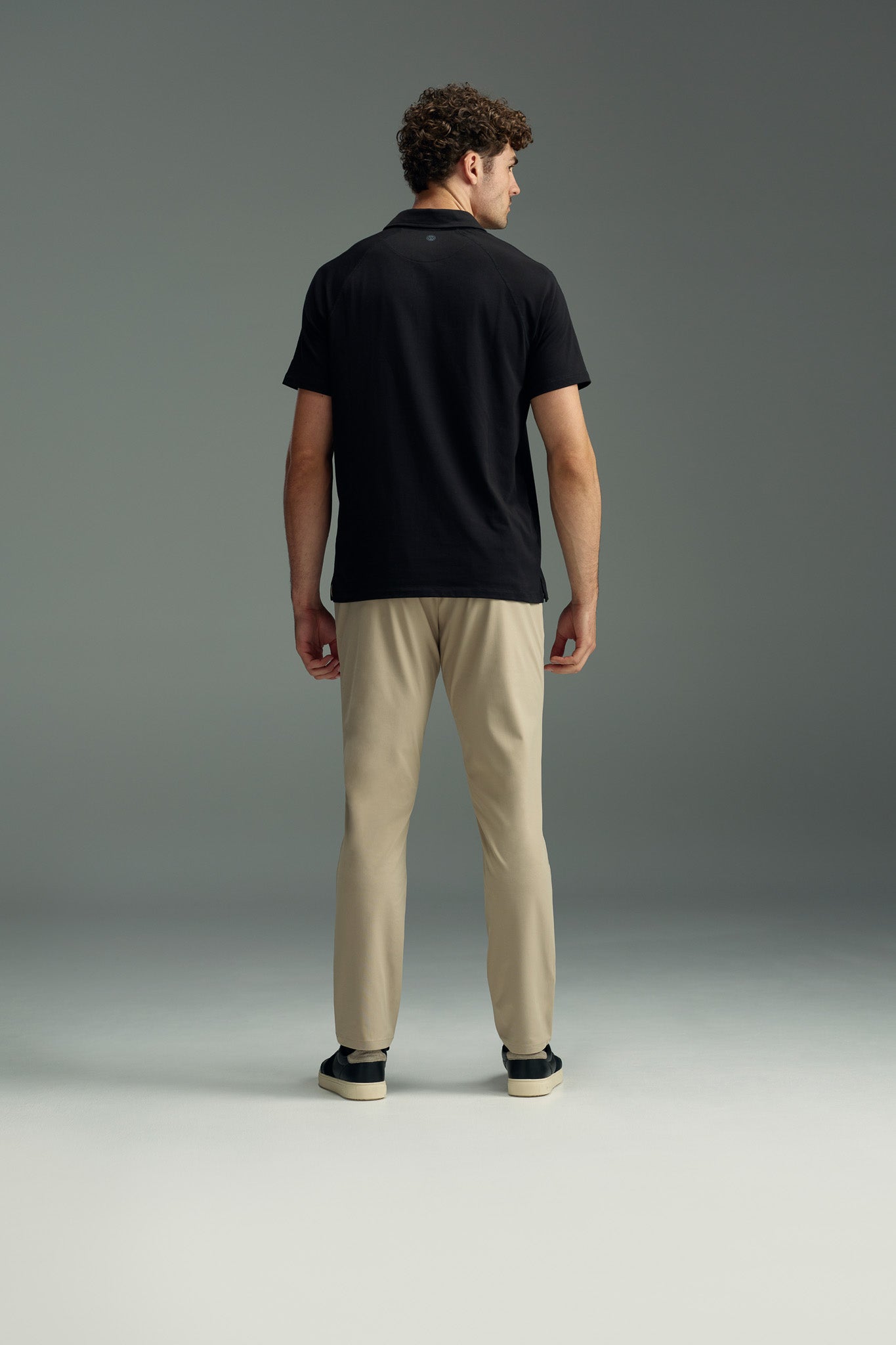 A man with short curly hair, dressed in a black polo shirt and WearEver Long Game Pant by WearEver, along with black sneakers, stands facing away from the camera against a plain gray background.