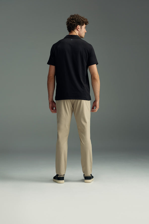 A man with short curly hair, dressed in a black polo shirt and WearEver Long Game Pant by WearEver, along with black sneakers, stands facing away from the camera against a plain gray background.