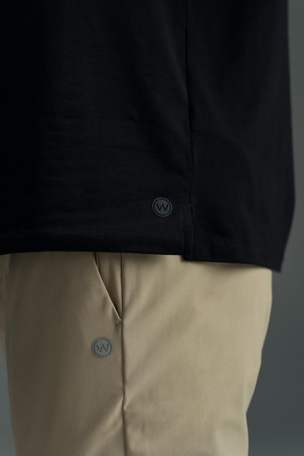 A close-up of someone wearing a black shirt and beige WearEver Long Game Pants, both marked with WearEver’s small circular WV logo near the hem and pocket.