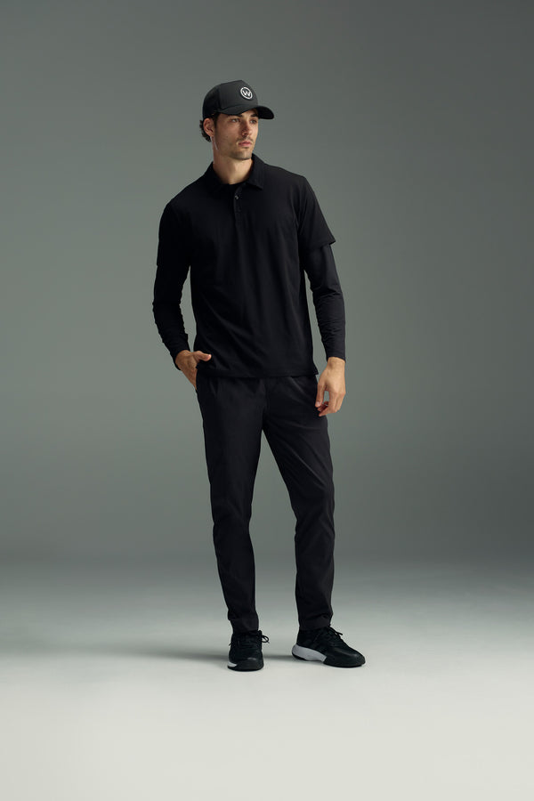 A man in a black baseball cap, black polo shirt, black long sleeves, WearEver Transit Pant by WearEver, and black sneakers stands with one hand in his pocket, looking off to the side against a plain gray background.