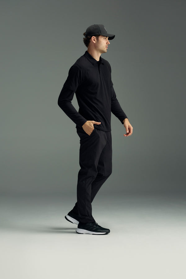 A man in profile wears a black baseball cap, a black long-sleeve polo, WearEver Transit Pant by WearEver, and black sneakers with white soles, standing against a plain gray background.