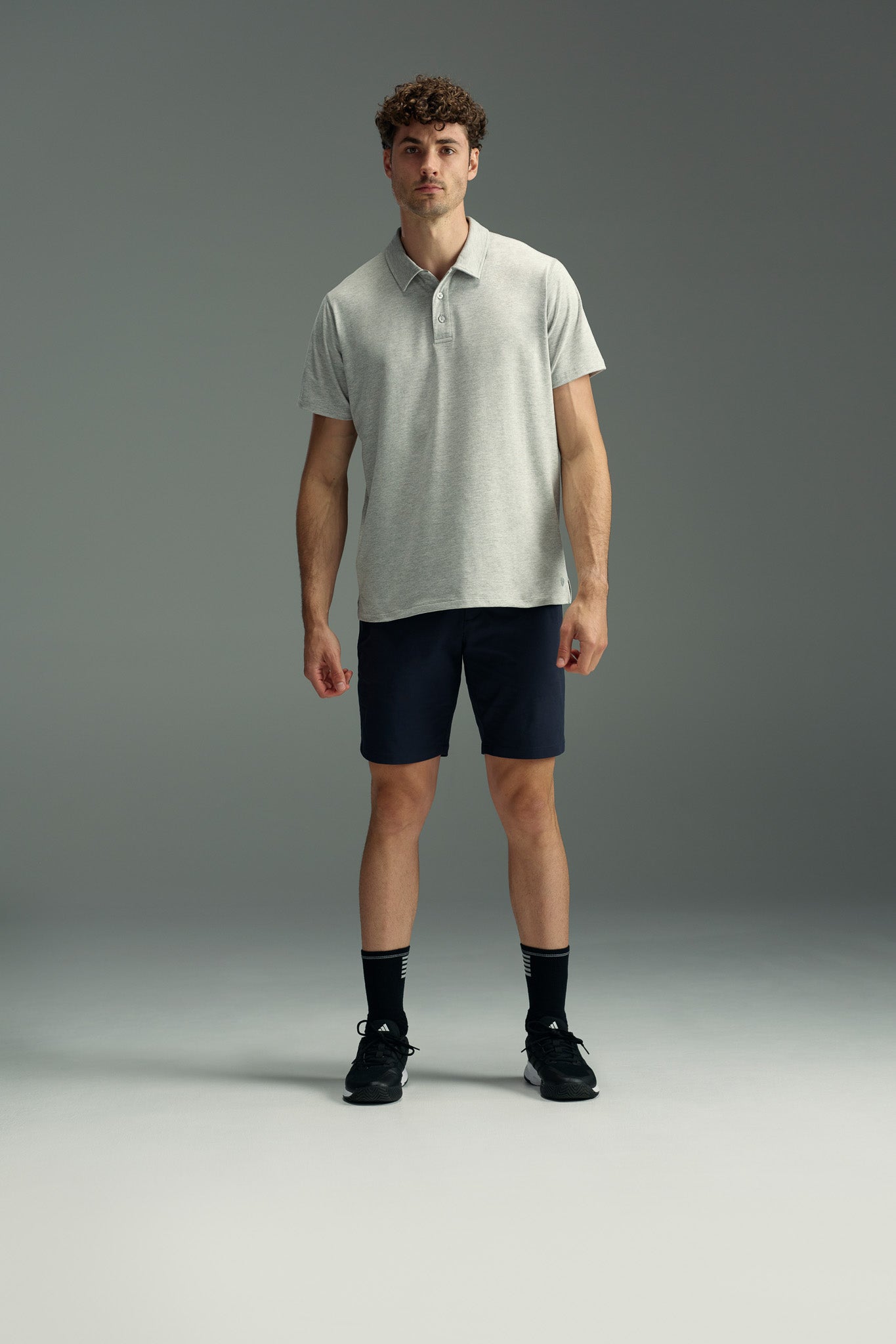 A man stands facing forward against a plain gray background, wearing a light gray WearEver Outbound Polo, dark navy shorts, black socks, and black sneakers.