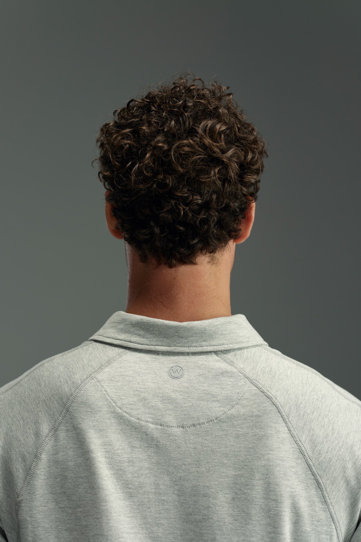 Seen from behind against a gray background, a person with short, dark, curly hair wears the WearEver Outbound Polo by WearEver—a light gray shirt crafted from quick-drying, breathable cotton-blend fabric.