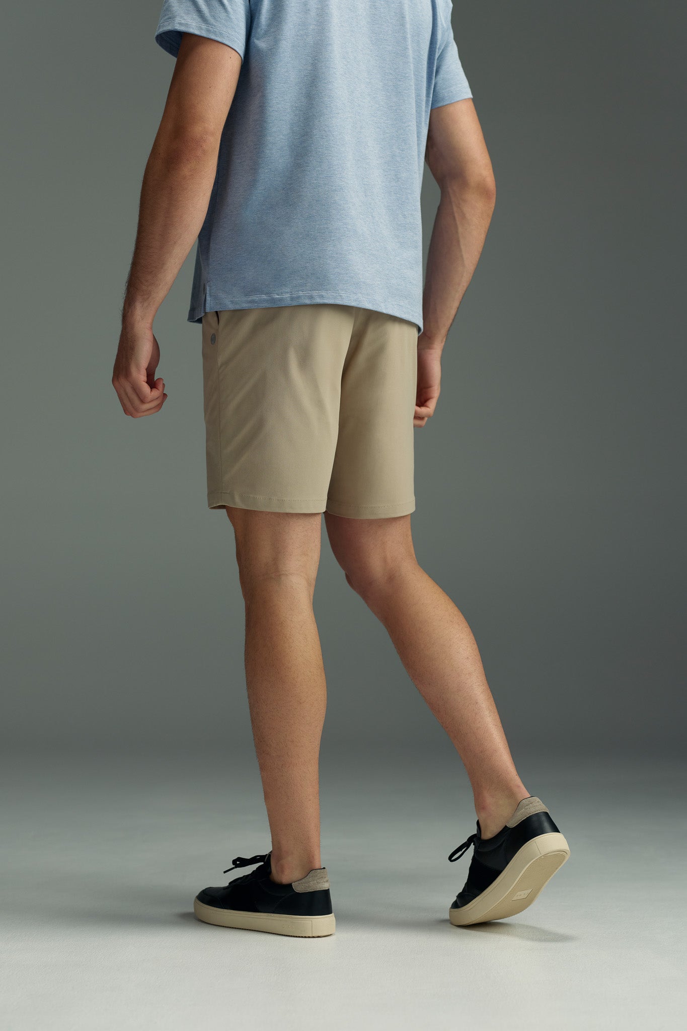 A person stands on a gray floor against a gray background, facing away from the camera, wearing a light blue t-shirt and WearEver Switch Shorts by WearEver in beige, paired with black sneakers featuring white soles.