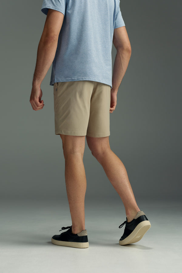 A person stands on a gray floor against a gray background, facing away from the camera, wearing a light blue t-shirt and WearEver Switch Shorts by WearEver in beige, paired with black sneakers featuring white soles.