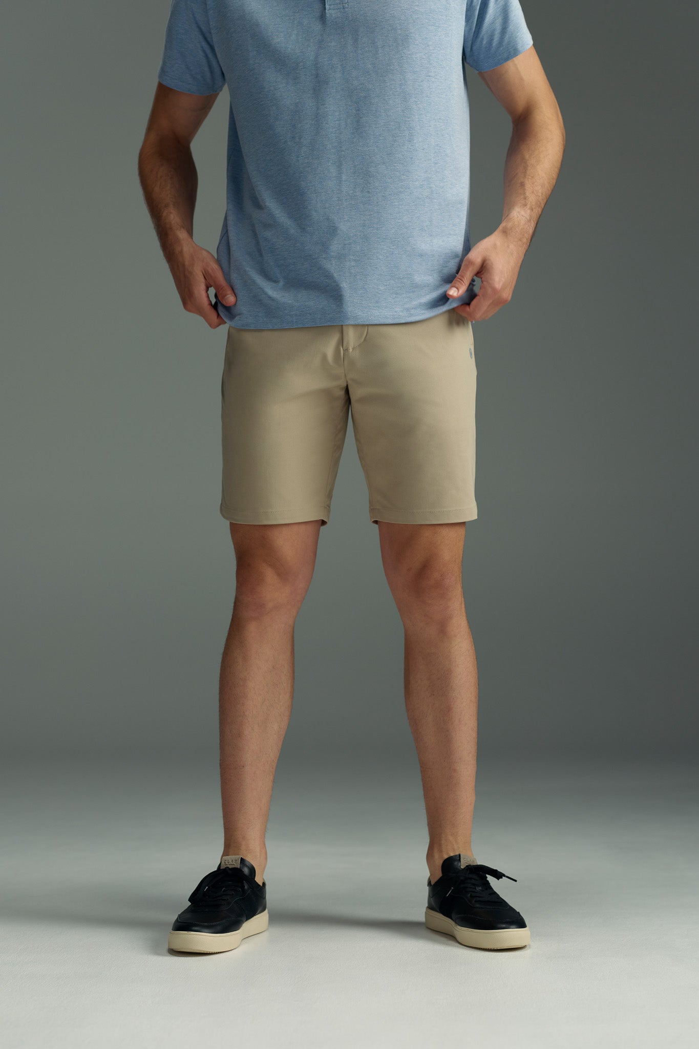 A person wearing a light blue short-sleeve shirt, black sneakers, and WearEver Switch Short by WearEver stands on a gray floor, holding the sides of their shorts. The image is cropped at the shoulders.