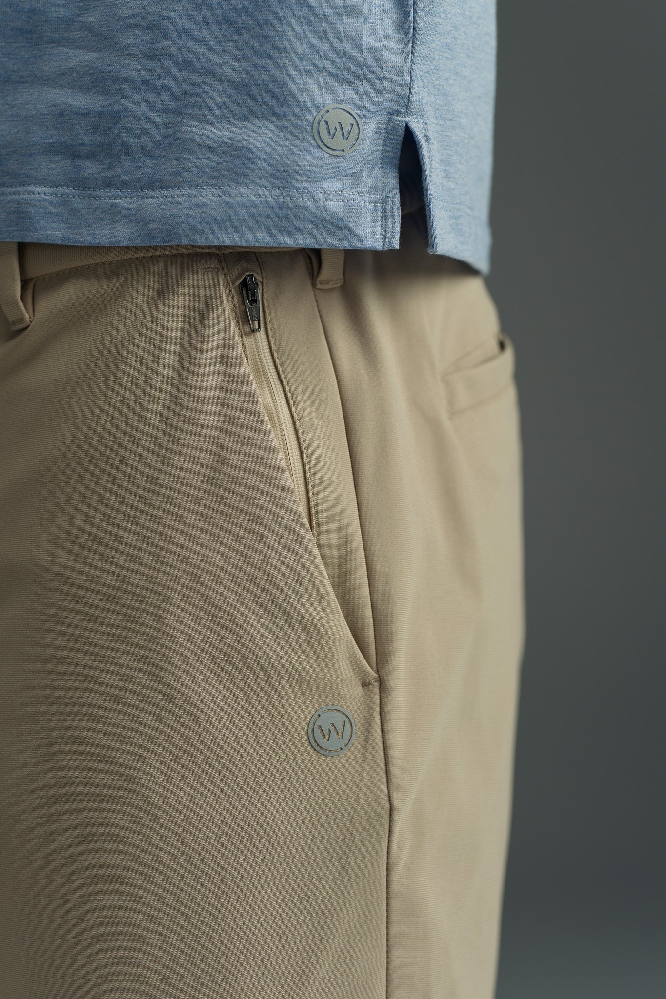 Close-up of a person wearing a light blue shirt and WearEver Switch Short by WearEver in beige with comfort stretch, featuring a visible zipper and small circular logos. The plain background is out of focus.