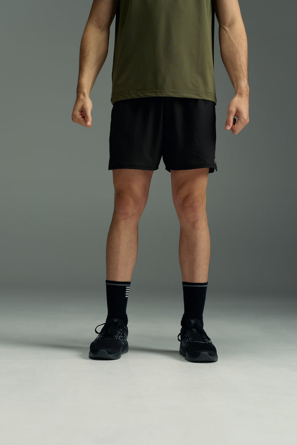 A person in a green t-shirt, WearEver Cadence Short in black, black socks, and lightweight breathable black athletic shoes stands against a gray background. The image is cropped at the shoulders, hiding the person’s face.